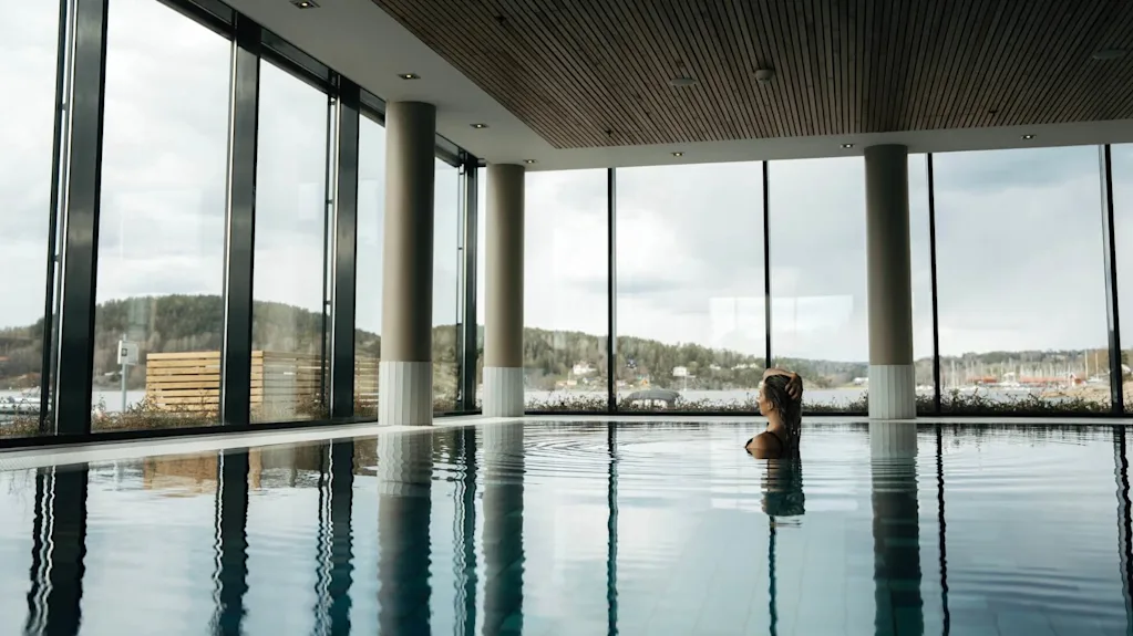 En person svømmer fredeligt i en indendørs pool omgivet af store vinduer med udsigt til træer og en overskyet himmel udenfor.