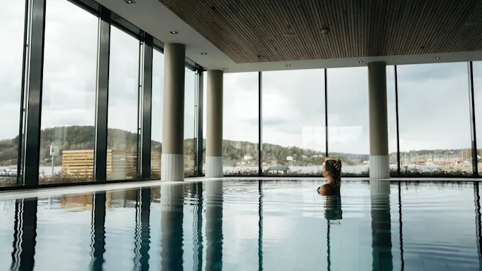 Person swimming peacefully in an indoor pool surrounded by large windows, with a scenic view of trees and a cloudy sky outside.
