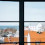 Udsigt gennem et vindue; orange-taget bygninger og parkerede biler i forgrunden. Træer og en stor vandmasse i det fjerne under en klar himmel.