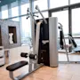 En multigym-maskin med trinser og sæder står ubrugt i et moderne fitnesscenter. Den er omgivet af pænt stablede håndklæder, mosaikvægsudsmykning og store vinduer.