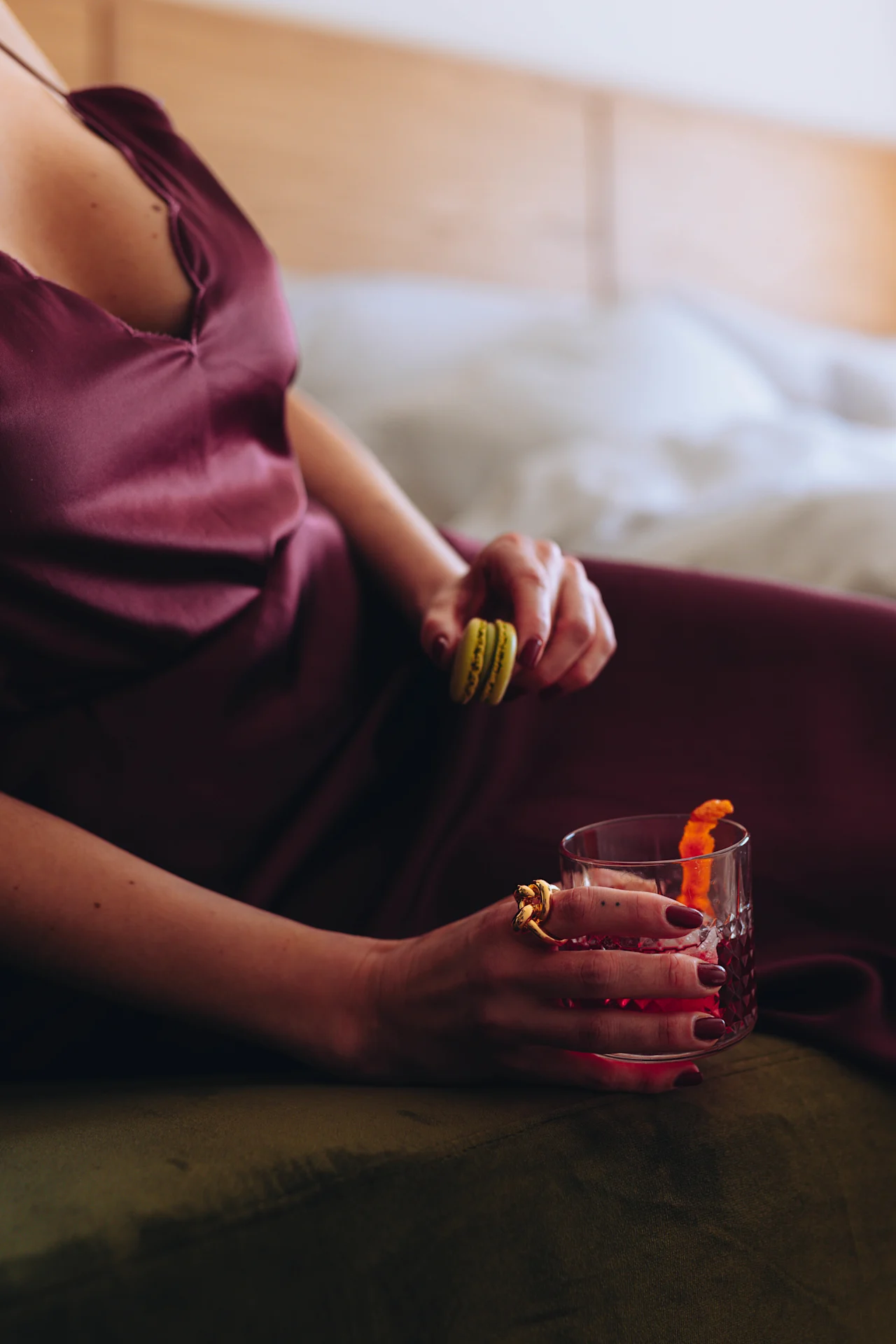 En person i en vinröd klänning håller en gul macaron och ett glas med röd dryck och apelsinskal, sittande på en säng med vita lakan.