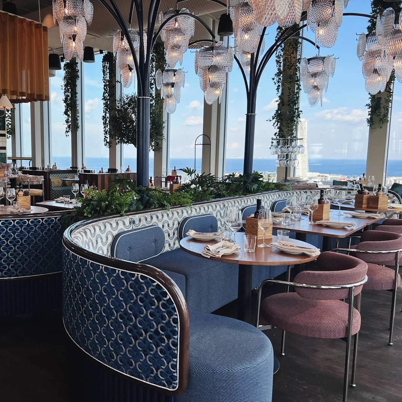 En stilfull restaurant har buede blå boder og trebord dekket for middag. Elegante lysarmaturer henger over, og store vinduer gir utsikt mot havet.