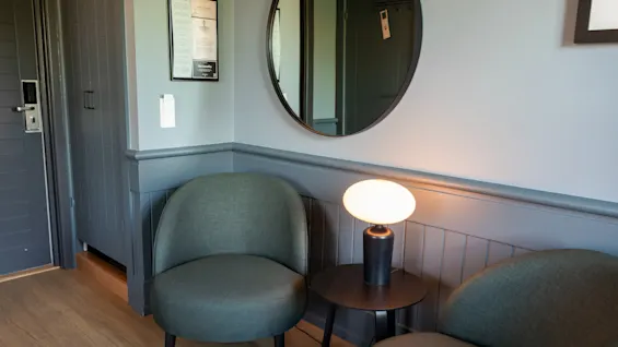 To grønne stole står over for et lille rundt bord med en tændt lampe. Et rundt spejl og en indrammet besked hænger på hvide vægge omgivet af træpaneler.