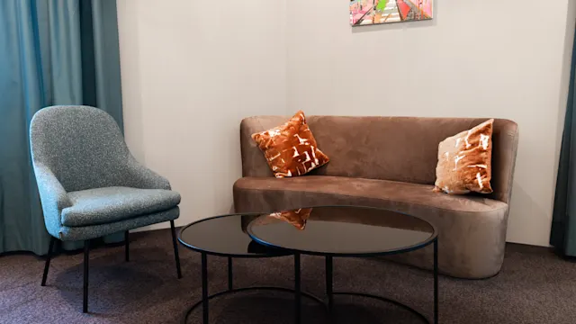 A small brown sofa with orange pillows sits beside a blue armchair, facing two black round coffee tables in a carpeted room. A colorful picture hangs on the wall.