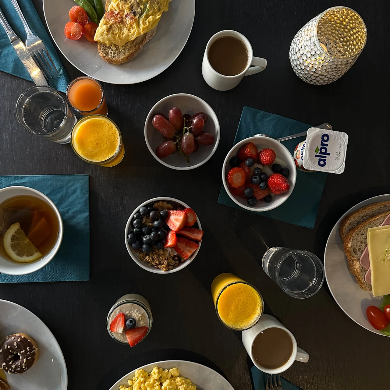 En varierad frukost uppställd på ett mörkt bord med ägg, frukt, bröd, drycker och yoghurt. Tallrikar och glas är ordnat med ett ljus för stämning. Text på yoghurt: "alpro."