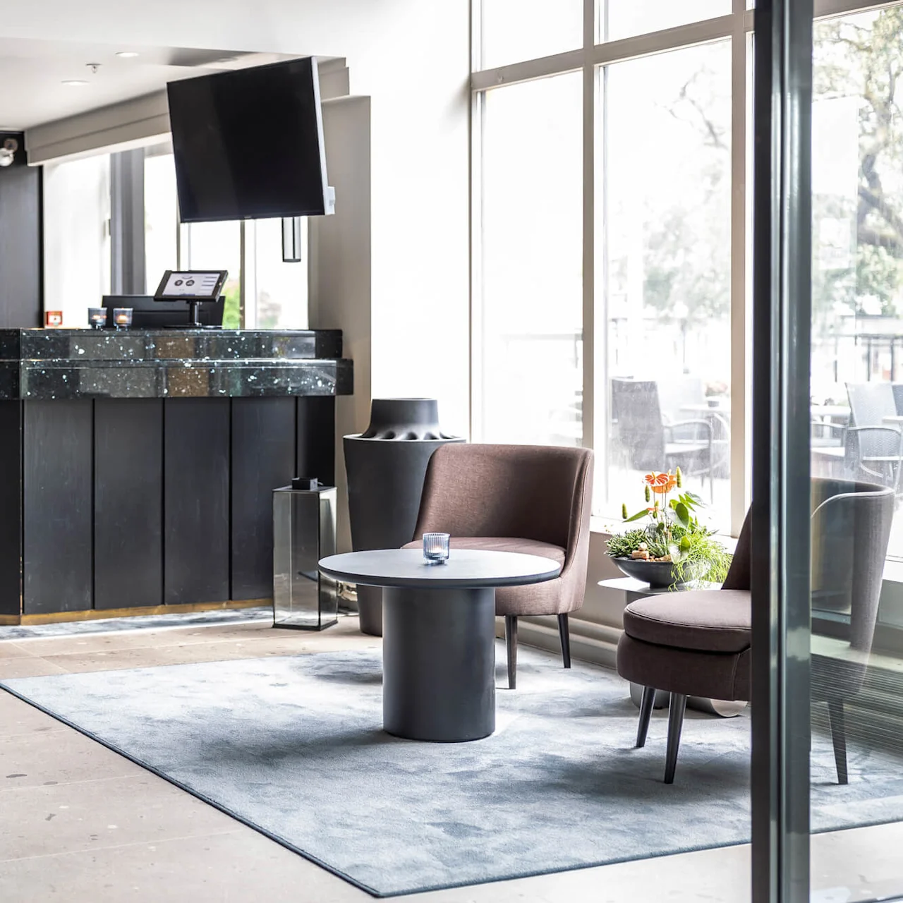 Reception med skiltet "QUALITY HOTEL Grand Larvik", mørk marmorskranke, tilstødende siddeområde med stole og bord, store vinduer oplyser rummet, delvis udsigt udenfor.