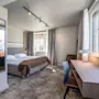 En pænt redt seng med brune og hvide lagener står ved store vinduer i et moderne, lyset hotelværelse. Et skrivebord med stol og lampe er i nærheden.