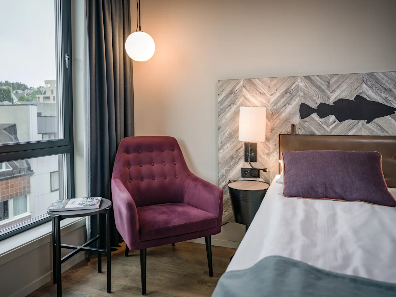 Moderni hotellihuone, jossa on mukava sänky, violetti nojatuoli ikkunan vieressä ja koristeellinen kalasiluetti sängynpäädyssä.