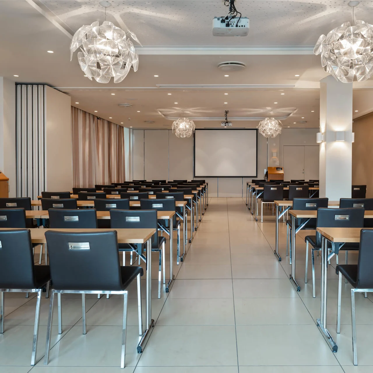 Klassrumsmiljö med rader av svarta stolar och träbord vända mot en vit projektorduk. Moderna ljusarmaturer hänger från taket. Väggarna är beige med minimalistisk inredning.