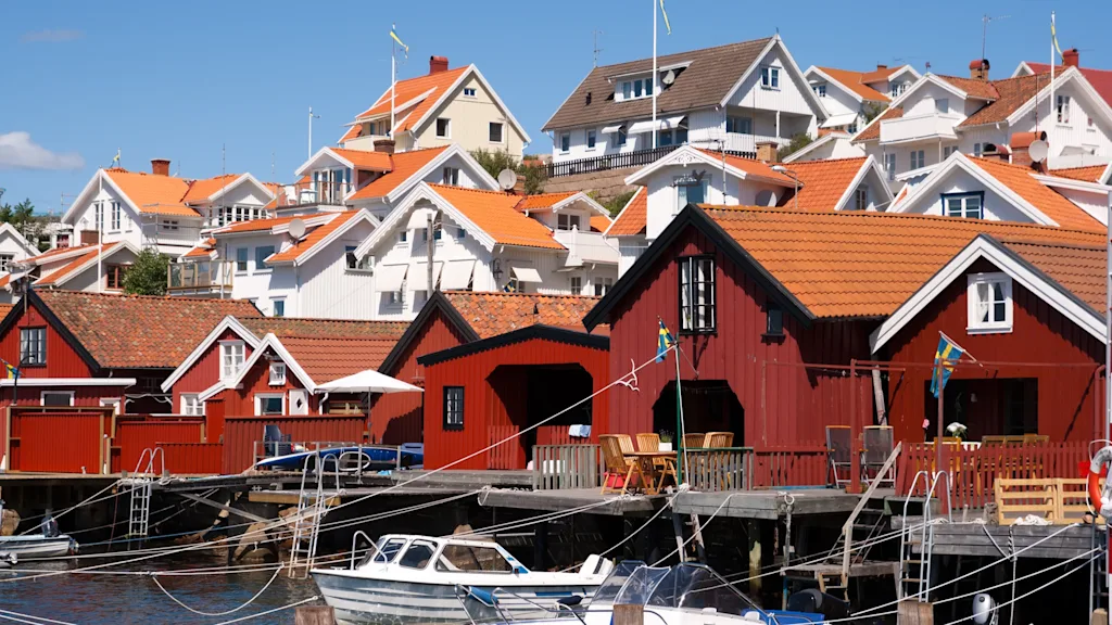 Röda träbåthus med bryggor ligger vid en lugn vattenfront. I bakgrunden reser sig vita hus med orangea tak på en kulle under en klarblå himmel.