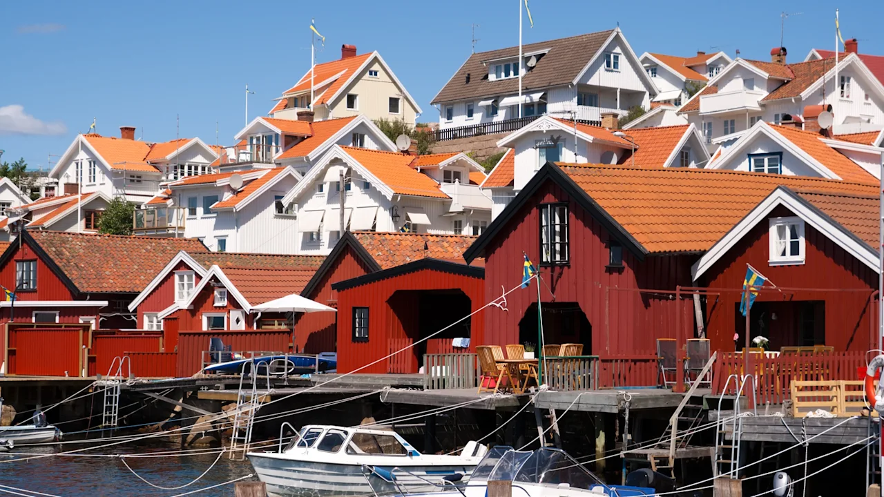 Røde træbådehuse med broer ved en stille vandfront. I baggrunden stiger hvide huse med orange tage på en bakke under en klarblå himmel.