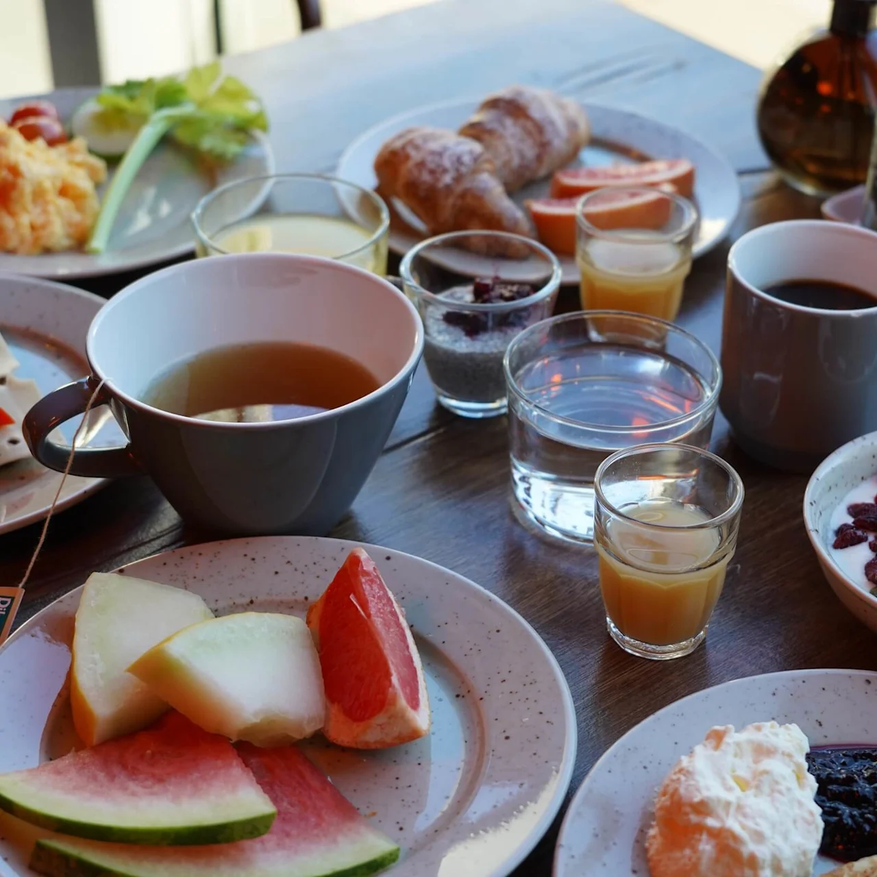 Assorted breakfast foods including fruit, yogurt with nuts, tea, coffee, juice, scrambled eggs, and pastries are arranged on a wooden table. Tea tag reads "Dilmah Exceptional."