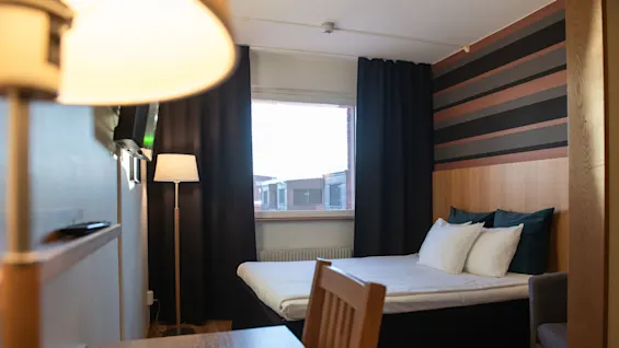 Bed positioned against a striped wall, with pillows neatly arranged. A soft chair and a wooden desk nearby. A floor lamp illuminates the room; window reveals buildings outside.