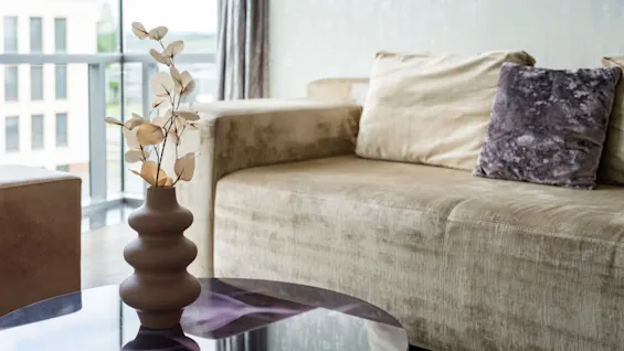 Brown vase with decorative white branches resting on a glass coffee table beside a beige sofa adorned with plush cushions. Large windows stream natural light into the modern living room.