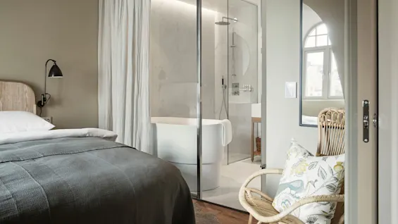 A bed with gray bedding sits beside a glass wall separating a modern bathroom with a freestanding tub and shower, set in a softly lit room with a wicker chair.