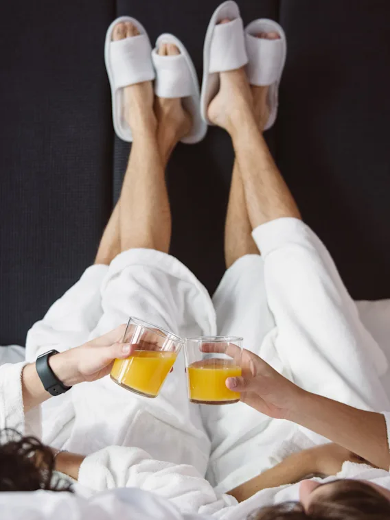 Två personer i badrockar och tofflor kopplar av i sängen och skålar med apelsinjuice. En mysig och glad stund av avkoppling.