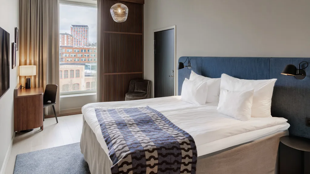 A modern hotel room with a neatly made bed, city view window, and stylish furnishings. The decor suggests an upscale urban accommodation.