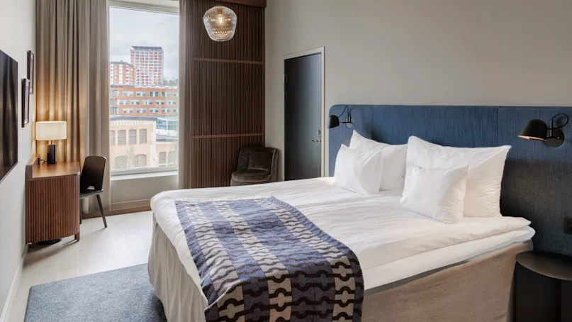 A modern hotel room with a neatly made bed, city view window, and stylish furnishings. The decor suggests an upscale urban accommodation.