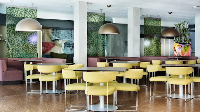 Yellow chairs are arranged around tables in a brightly lit, modern dining area with decorative green panels and large hanging lamps.