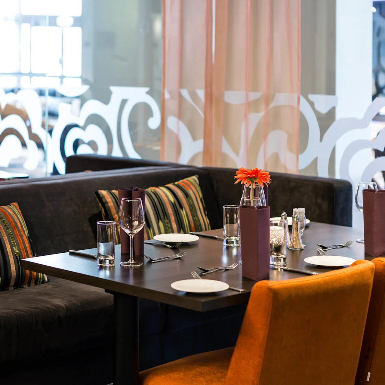 A dining table is set with glassware and orange chairs in a stylish restaurant, decorated with striped cushions and a floral centerpiece amid softly lit surroundings.