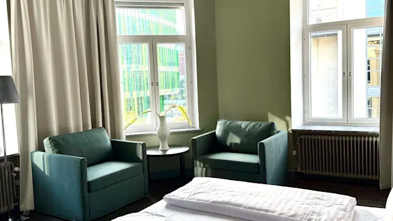 Ett mysigt hotellrum med en säng, två fåtöljer och stora fönster, i en lugnande grön färgskala.