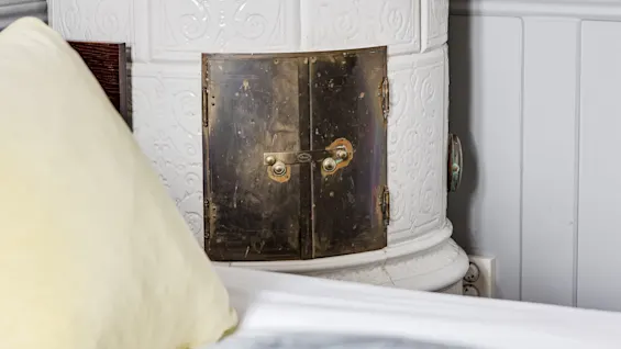 A white ceramic stove with an ornate design features a brass door. It stands beside a bed with a yellow pillow, against a gray wall with paneling.
