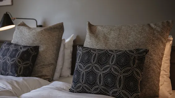 En bekväm säng prydd med en mängd texturerade och mönstrade kuddar, mjukt upplyst av en sänglampa, vilket skapar en mysig atmosfär.