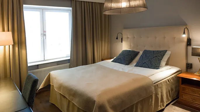 En seng med to blåmønstrede puter hviler under en stor lampe, omgitt av beige gardiner, et vindu og et skrivebord med en stol i et koselig rom.