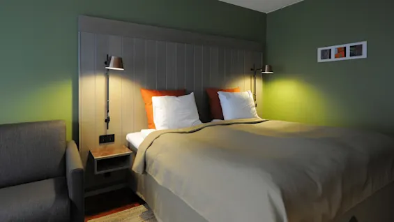 A neatly made bed with white and orange pillows rests against a wood-paneled wall. Overhead lamps provide light in a room with green walls and a gray sofa.