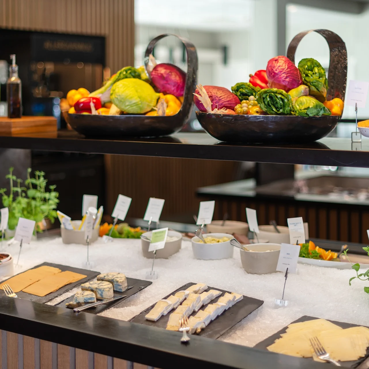 Two large bowls of colorful vegetables sit atop a buffet counter, surrounded by assorted cheeses, condiments, and herbs, displayed with small signs. 