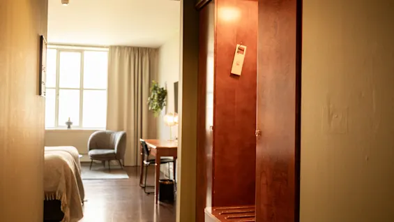 Et træskab med et papirmærke hænger åbent i hotelgangen, der fører til et soveværelse med seng, skrivebord og vindue.