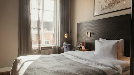 Ett solbelyst hotellrum på Home Hotel Grand, Helsingborg, med en bekväm säng och ett skrivbord vid fönstret. Text: MIT LIV.