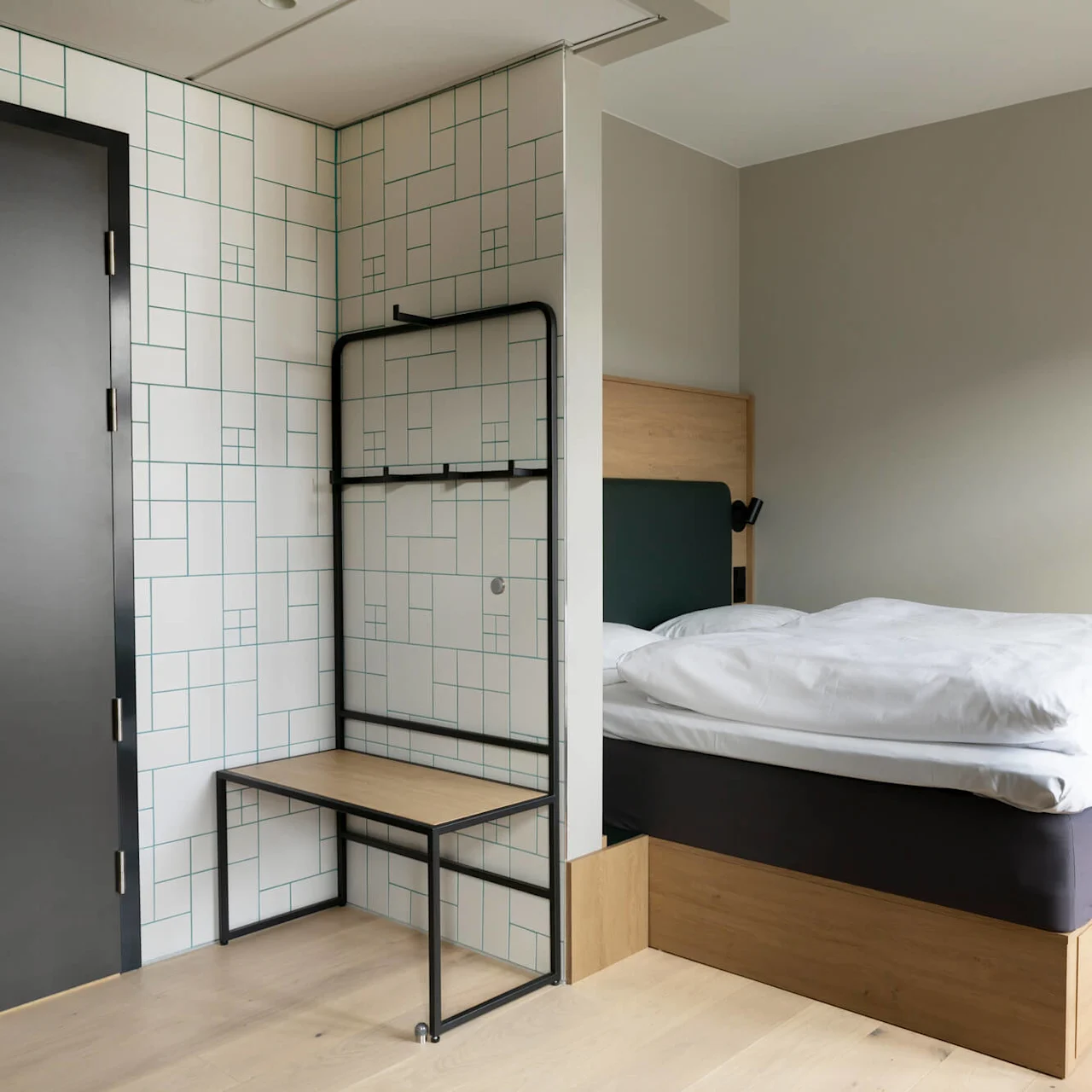 A bed with white linens stands in a minimalist room, next to a patterned wall panel and a black door, creating a contemporary, airy atmosphere.