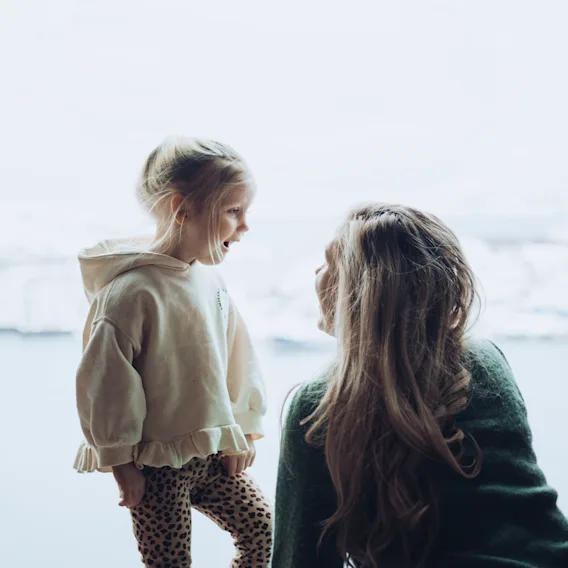 An adult and a child by a large window, looking out and sharing a moment of observation or conversation.