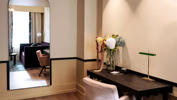 A decorative arched mirror reflects a room with a purple sofa and table, while nearby, a black desk with a green lamp is adorned with a vase of colorful flowers.