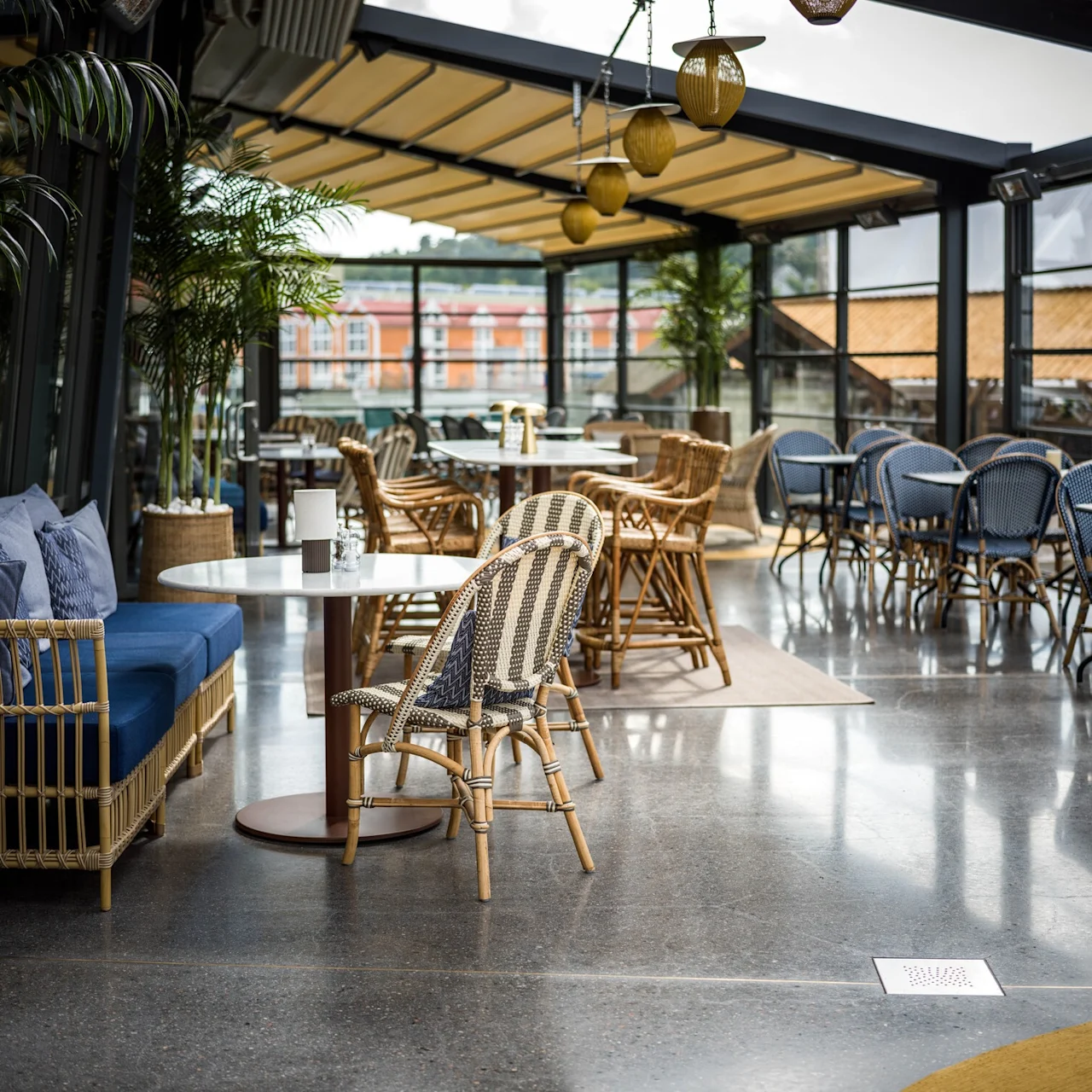 Café sittgrupp med rottingstolar och bord på ett polerat betonggolv, omgivet av stora glasfönster. Krukväxter ger grönska. Texten "SOCIAL" synlig på glaset.