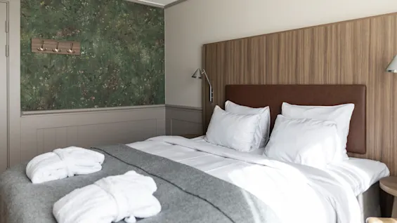 Viihtyisä hotellihuone, jossa on pedattu sänky valkoisilla liinavaatteilla ja harmaalla peitolla, sekä kaksi kylpytakkia.