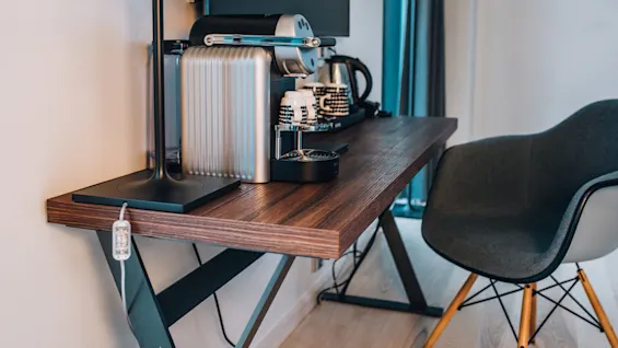 En kaffemaskine står på et træbord nær en lampe, kopper og en kedel i et moderne rum. En stol er i nærheden.