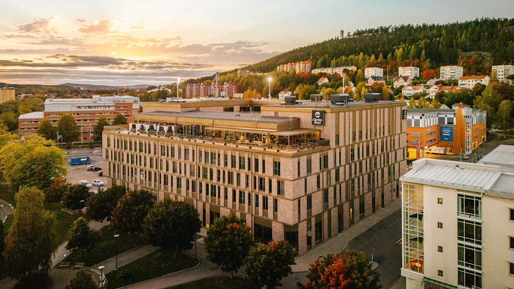 En stor tegelbyggnad med "Clarion Hotel"-skylt ligger i en stadsmiljö omgiven av träd och andra byggnader under solnedgången. En sluttning med hus syns i bakgrunden.