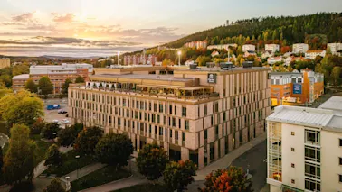 A large brick building with "Clarion Hotel" signage is set in a cityscape surrounded by trees and other buildings during sunset. A hillside with houses is visible in the background.