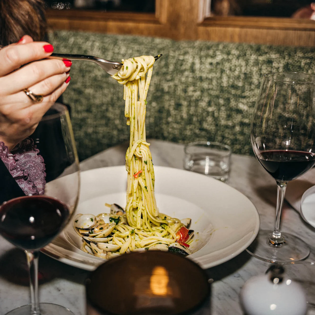 A person twirls spaghetti with clams on a fork, accompanied by two glasses of red wine, at a restaurant table with a green cushioned bench.
