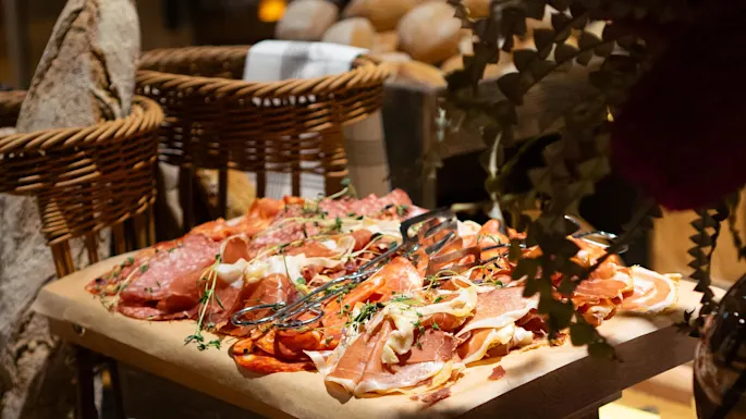 Cured meats on parchment paper atop wooden crates, surrounded by bread baskets and greenery in a rustic setting. Sign reads: "PEPPARSALAMI PEPPERSALAMI".