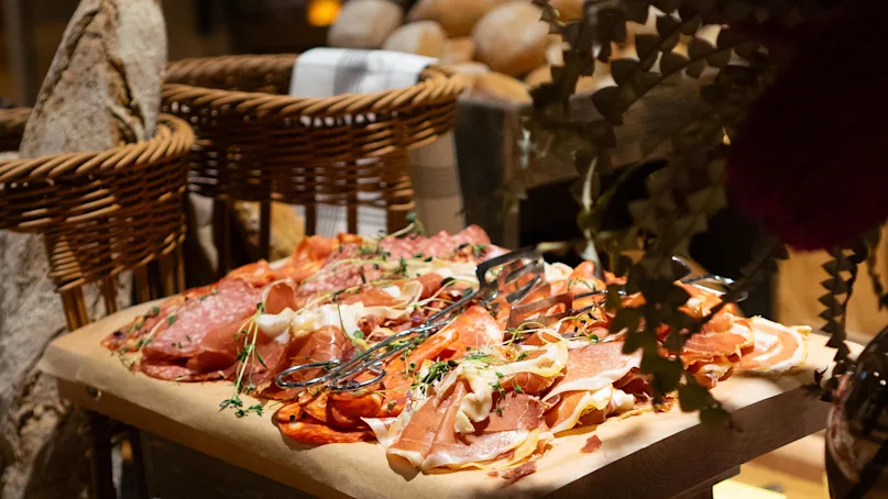 Cured meats on parchment paper atop wooden crates, surrounded by bread baskets and greenery in a rustic setting. Sign reads: "PEPPARSALAMI PEPPERSALAMI".