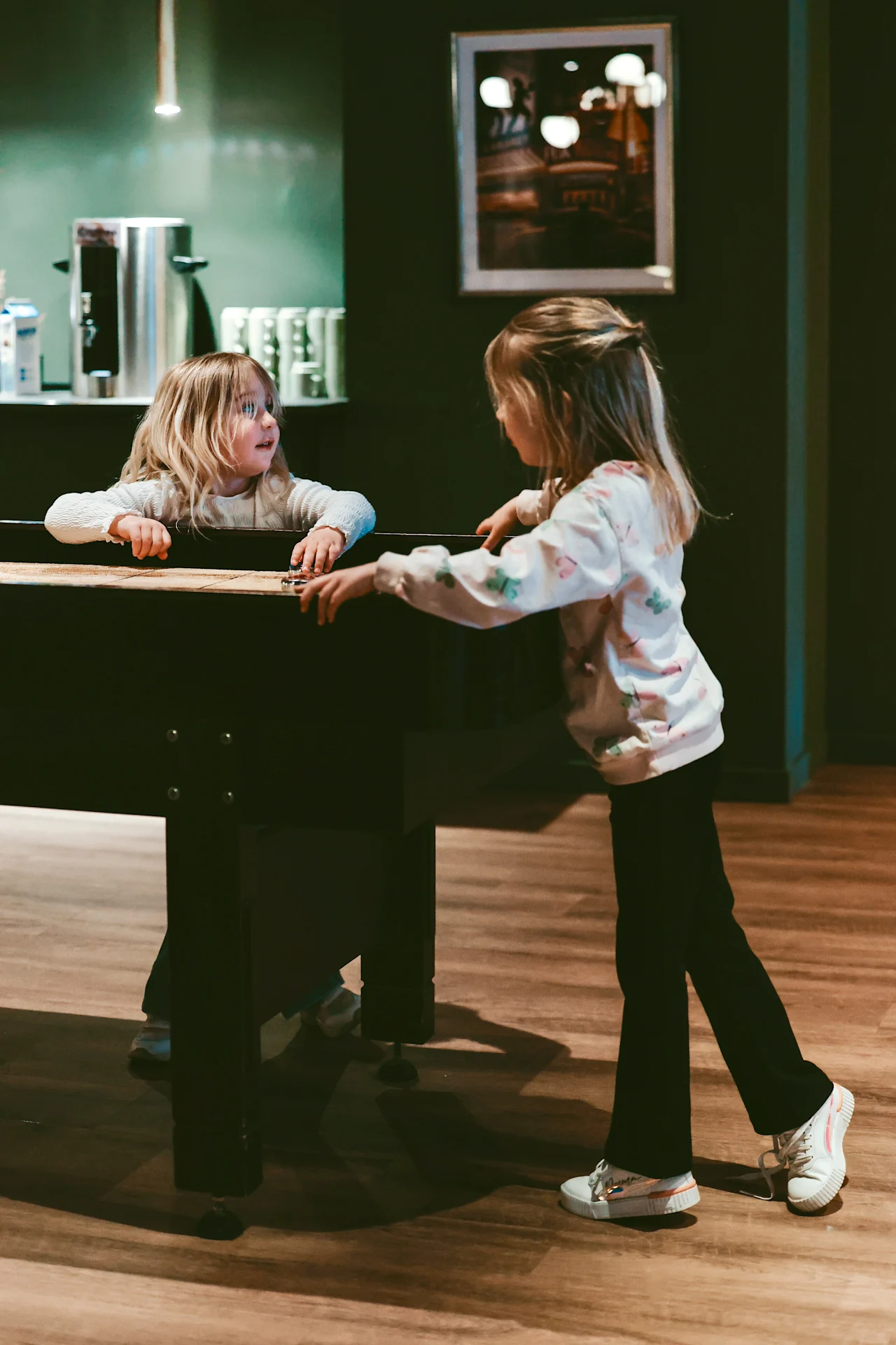Children interact by a foosball table in a cozy room with wooden flooring. A shelf holds mugs and a pot. A framed picture hangs on the wall.