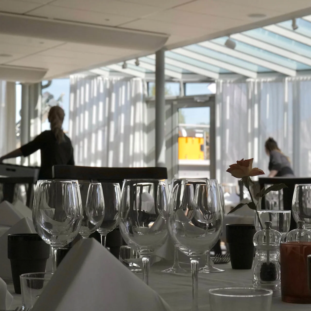 Borde med glas og servietter er pænt anrettet i en lys restaurant, mens to personer forbereder sig i baggrunden under et glasloft.