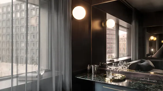 Et mørkt moderne hotellrom med en marmortoppet bardel som har glass og en isbøtte. Store vinduer med lette gardiner avslører en bysilhuett med høye bygninger.