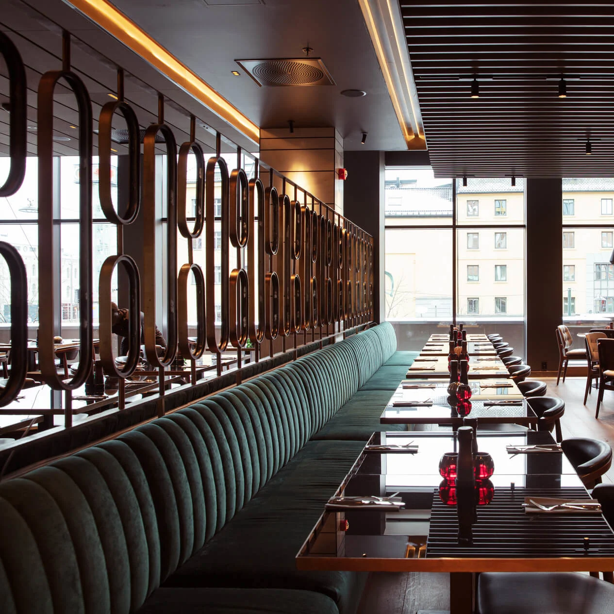 Elegant restaurant with long benches and tables, set with folded napkins and red glass candle holders. Large windows show a cityscape, and hanging pendant lights provide warm lighting.
