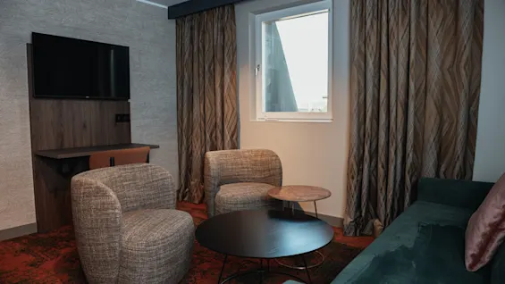 Et koselig sitteområde på hotellrommet med en TV, to komfortable lenestoler, en sofa og et vindu med gardiner.