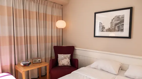 En seng står pænt i et hyggeligt rum med en vinrød stol, et lille træbord og en lampe. Et indrammet sort-hvidt fotografi hænger på den beige væg.