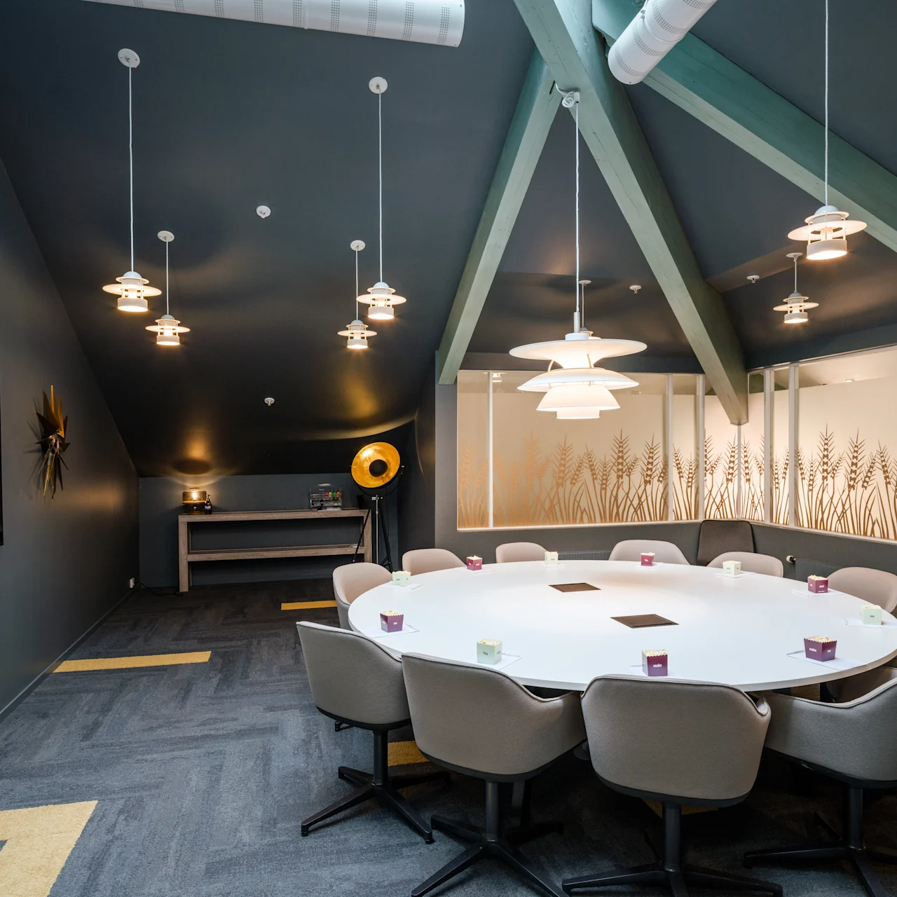 Round table surrounded by chairs, positioned under hanging lamps, in a modern conference room with a decorative wall featuring plant graphics.